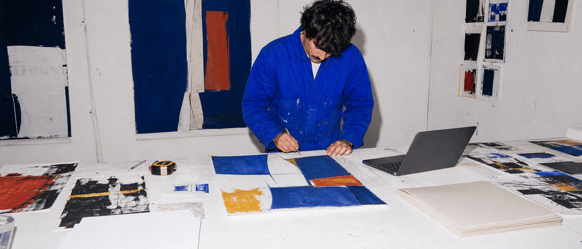 Artist working at a studio table beside a laptop and prints